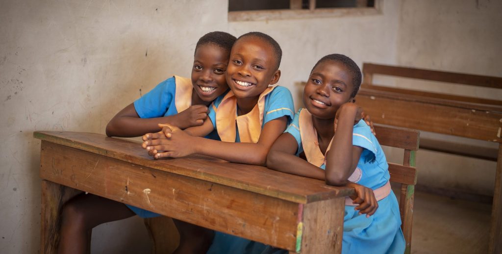 Three cheerful schoolgirls in blue uniforms smiling at the camera while sitting at a wooden desk in a classroom, showcasing a positive learning environment.