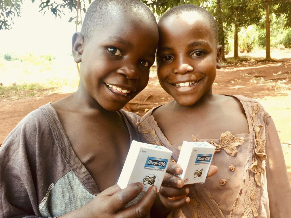 Two smiling children hold boxes of Alzol-400 in a rural outdoor setting, showcasing a moment of joy and access to essential medication.