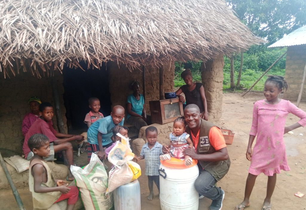 Group of children and adults gathered outside a rural mud house with a thatched roof, showcasing community life. Several children are playing while an adult smiles and holds a toddler on a container, with bags of food supplies in the foreground.