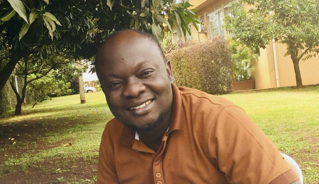 Smiling man wearing a brown polo shirt sitting on grass in a garden, surrounded by trees and a house in the background.