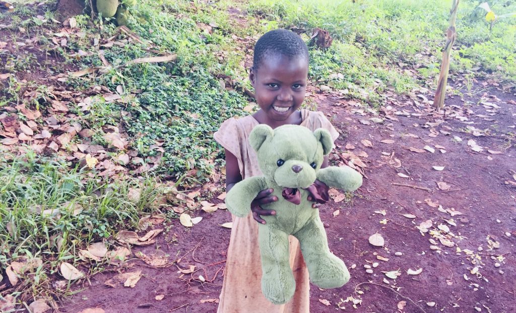 Smiling girl holding a green teddy bear in a natural outdoor setting with fallen leaves and grass. The child is wearing a simple dress and appears joyful, showcasing the bond between children and their toys.