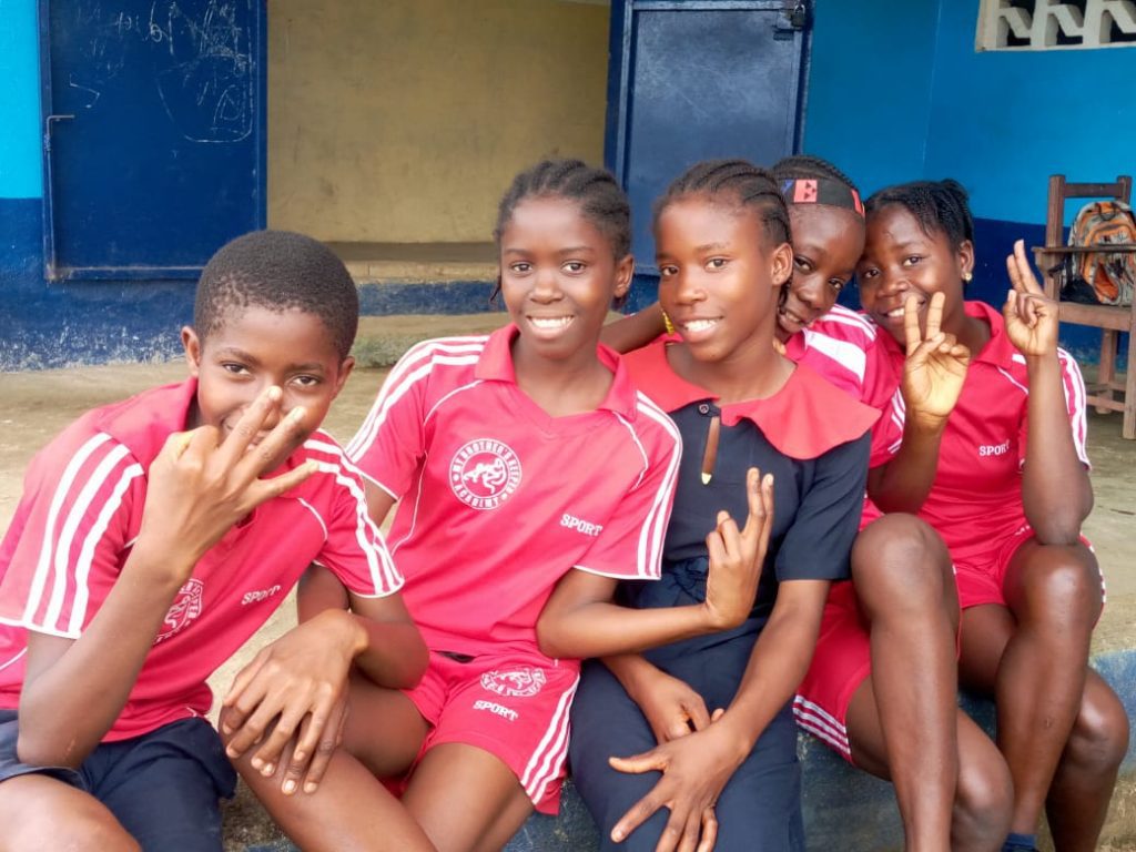 Group of five young girls smiling and posing for a photo, wearing matching red sports uniforms, outdoors in a school setting. The background features a blue wall and a door, emphasizing a vibrant and playful atmosphere.