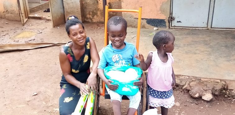 A woman and two children sitting outside a building in a rural area, with the woman holding shopping bags and the boy smiling while holding a container of food. The scene captures family bonding and community life.