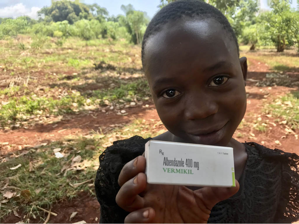 Smiling child holding a box of Albendazole 400 mg Vermikil in a rural setting, highlighting the importance of medication for treating parasitic infections.