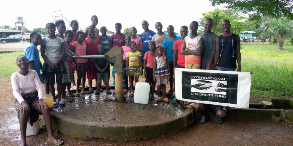 Group of children and adults gathered around a water pump, smiling and holding a banner for the International Children's Fund, highlighting community engagement and access to clean water.