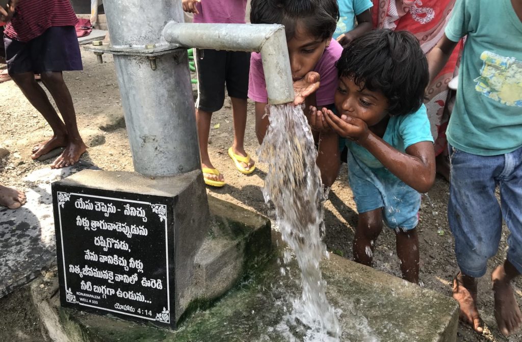 Children joyfully drinking water from a hand pump in a rural area, with a sign in the foreground highlighting the well's importance. The scene captures the essence of community access to clean water and the joy it brings to kids.