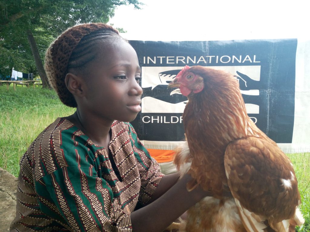 A young girl in traditional attire gently holds a brown chicken, showcasing her connection with animals at an event organized by the International Children’s Fund. The background features greenery and a banner promoting children's welfare.