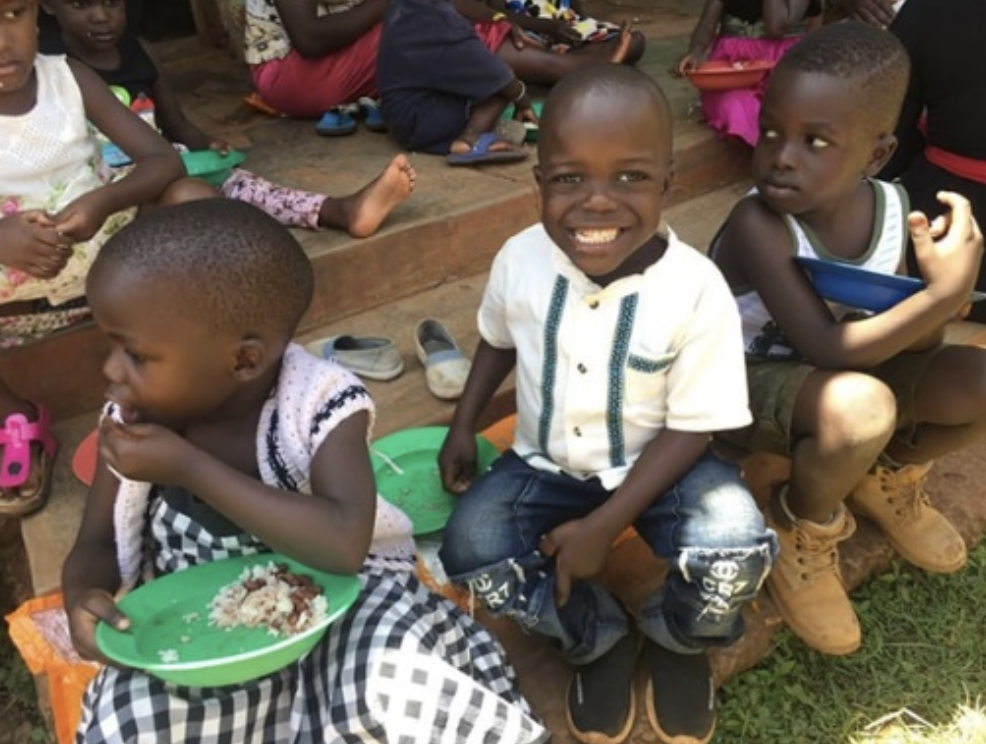 Children enjoying a meal outdoors, with one boy smiling widely while wearing a white shirt and blue jeans, a girl in a black and white dress eating, and another boy sitting beside them looking on. The scene captures a moment of joy and community among the kids.