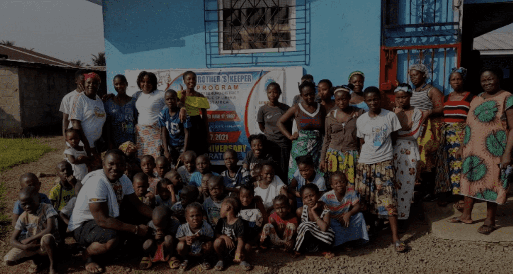 Group photo of community members, including children and adults, gathered outside a blue building for the Brother's Keeper program anniversary celebration in Liberia, showcasing local culture and community engagement.