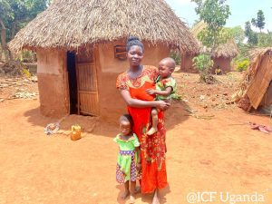 A mother holding a baby stands with her young daughter in front of traditional Ugandan mud huts, showcasing rural family life in Uganda. The scene features a red earth background and thatched roofs, highlighting the cultural setting.