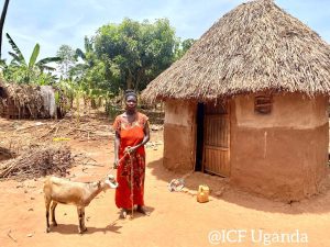 Woman in a red dress standing with a goat in a rural Ugandan village, featuring traditional mud hut and lush greenery in the background.