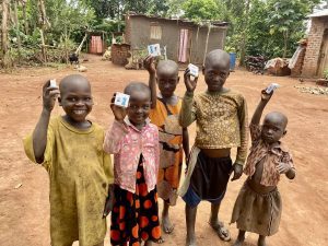 Children in a rural village proudly hold up small boxes, smiling for the camera. The background features traditional homes and lush greenery, highlighting their joyful expressions and community setting.