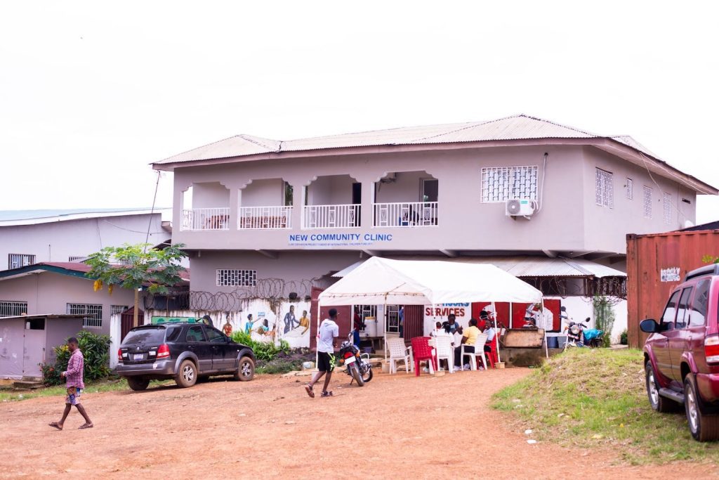 Exterior view of the New Community Clinic, featuring a two-story building with a balcony, surrounded by vehicles and people. The clinic serves as a healthcare facility in the community, with a service tent visible in the foreground.