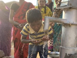 Young boy collecting water from a hand pump, demonstrating access to clean water in a rural community, with women in colorful traditional attire in the background.