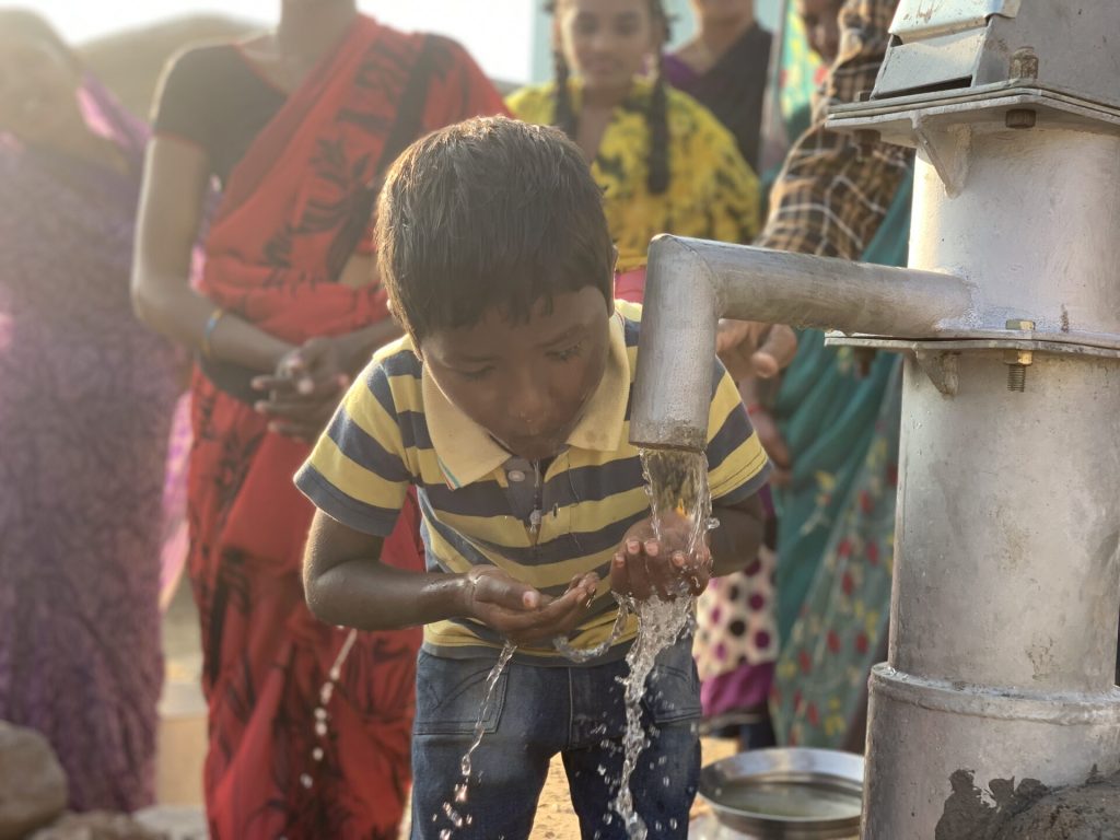 Child drinking water from a hand pump, surrounded by adults in traditional attire, highlighting access to clean water in rural communities.