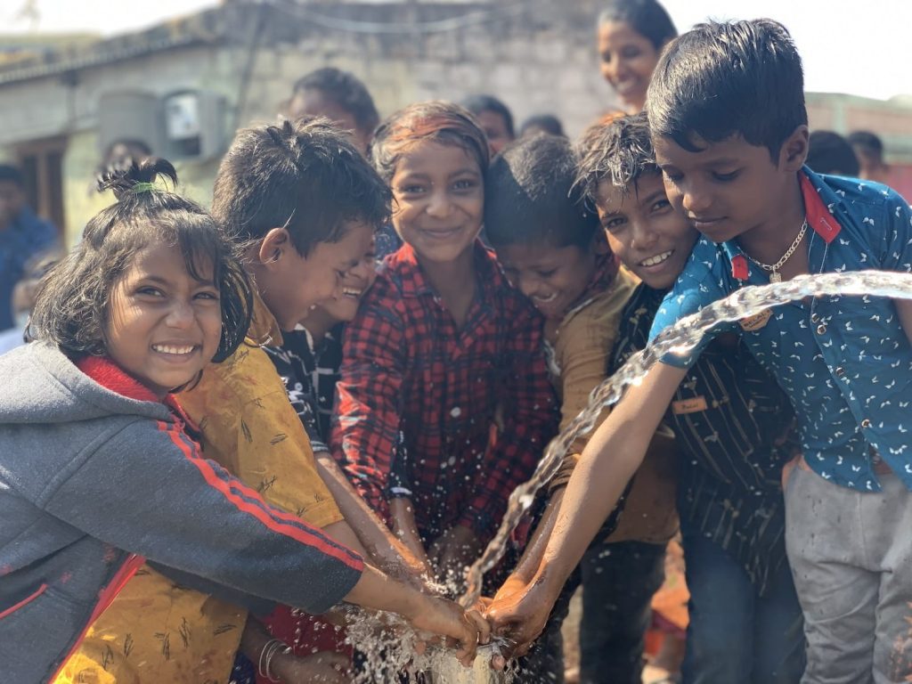 A group of joyful children playing with water, smiling and laughing, while standing together in a sunny outdoor setting. The scene captures the essence of childhood joy and community, highlighting their interactions and playful spirit.