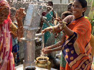 Women joyfully drawing water from a hand pump, splashing water around, showcasing community engagement and access to clean water in a rural setting.