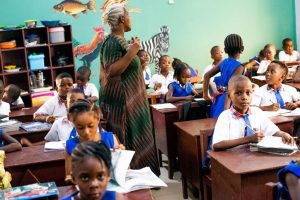 Classroom scene with diverse children in blue uniforms attentively listening to a teacher. The vibrant background features animal murals, and students are engaged with their notebooks and textbooks, highlighting an interactive learning environment.