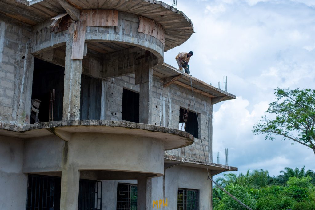 Construction worker on scaffolding performing tasks on a multi-story building under construction, showcasing concrete structure and unfinished exterior. Lush greenery in the background with a partly cloudy sky.