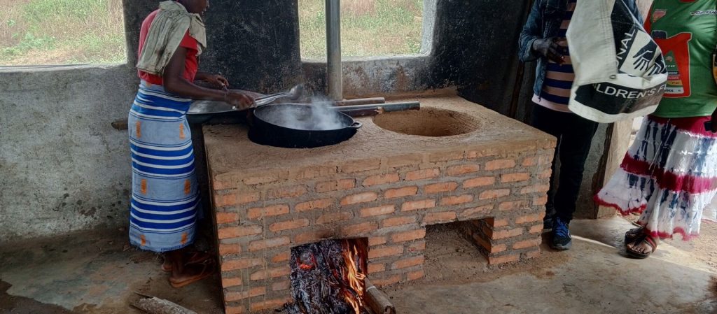 Woman cooking on a traditional brick stove in a rustic kitchen, with smoke rising from a large pot. The scene depicts a communal cooking environment, showcasing local culinary practices and the use of a wood fire for heat.