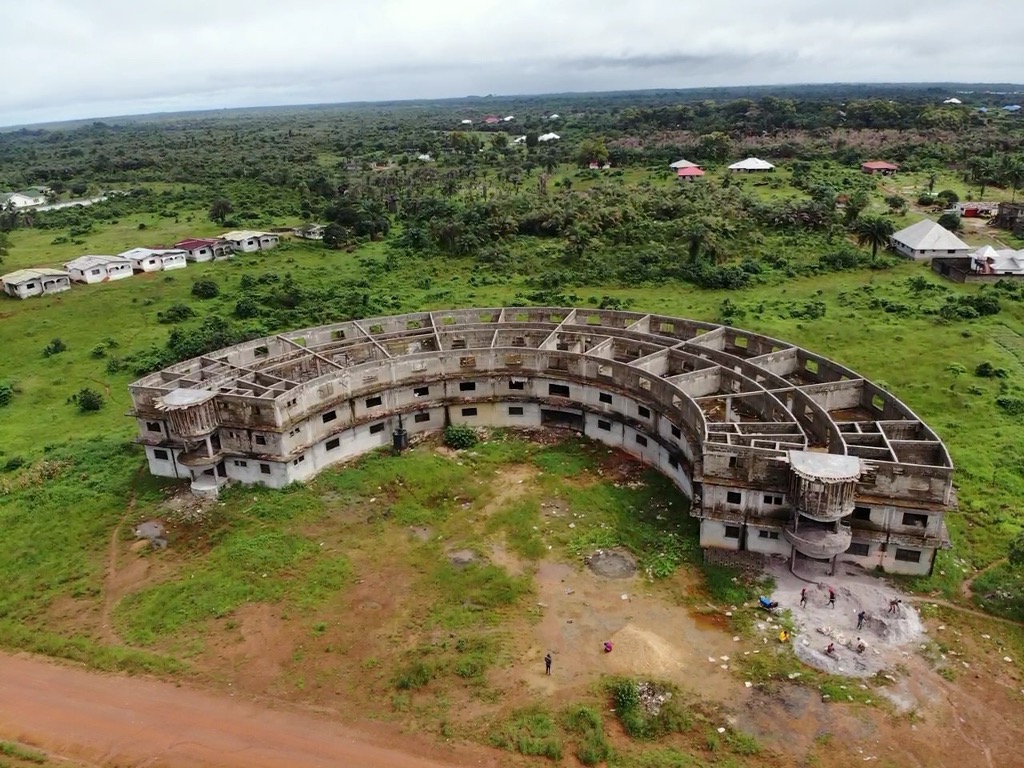 Aerial view of an abandoned, partially constructed building surrounded by lush greenery and scattered residential structures, highlighting the contrast between urban decay and natural landscape.
