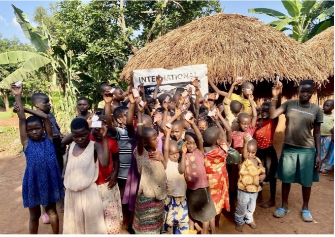 A group of children and adults in a rural setting, gathered in front of a thatched-roof hut, holding up cards in celebration. The scene reflects community engagement and support from an international organization, surrounded by lush greenery and a clear blue sky.