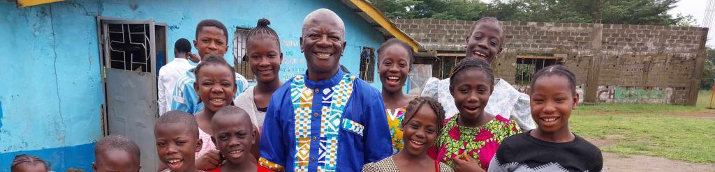 Group of smiling children and an adult man in colorful traditional attire, standing outside a blue building, showcasing joy and community spirit in a vibrant setting.