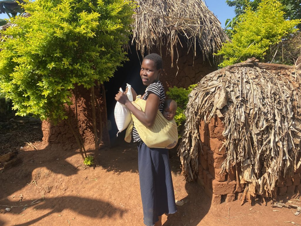 A woman holding a bag of rice while carrying a baby on her back, standing in front of a traditional mud house surrounded by greenery in a rural setting.