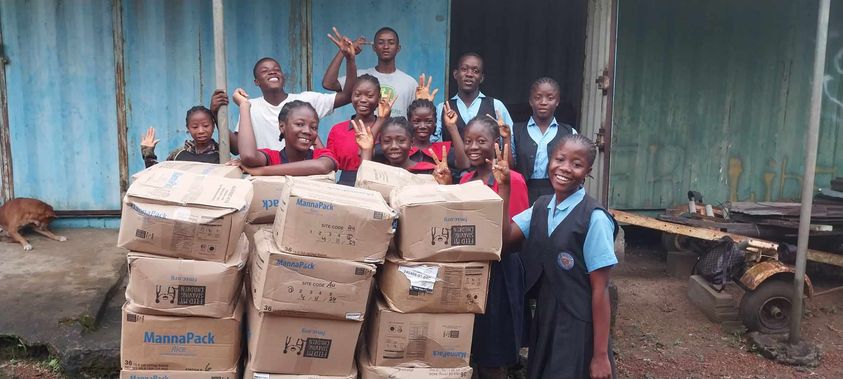 Group of children and teenagers smiling and posing with boxes of MannaPack food donations outside a building, promoting community support and hunger relief efforts.